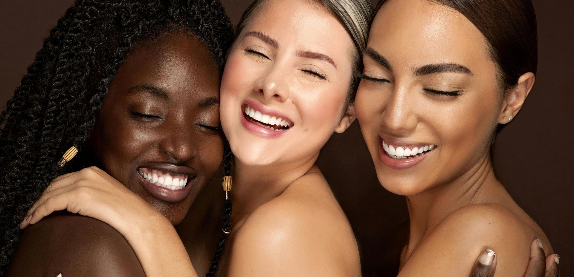 Three diverse women embracing with eyes closed and smiling, celebrating friendship and unity.