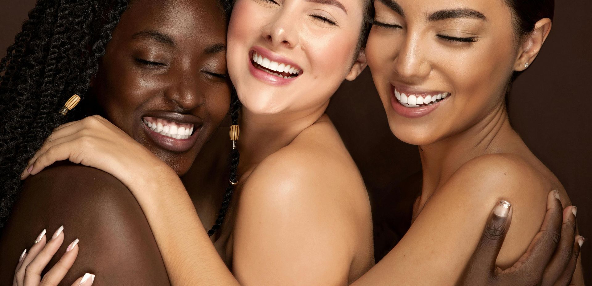 Three diverse women embracing with eyes closed and smiling, celebrating friendship and unity.